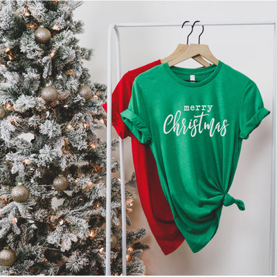 Green tee with white merry Christmas on front of tee hanging on a rack in front of tree