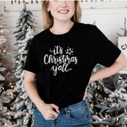 Woman wearing black tee with with text that reads it's Christmas y'all with a start in front of Christmas trees 