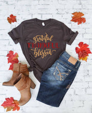 brown short sleeve Grateful Thankful Blessed T-Shirt tied on the side in a knot laid flat with folded jeans to right and brown boots to lower left fall leaves scattered and white and grey brick background