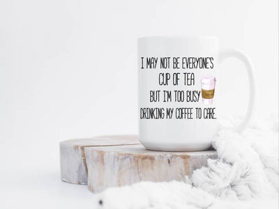 white ceramic coffee mug with handle to the right i may not be everyone's cup of tea but i'm too busy to care printed on front mug sitting on white wash slab of wood with a blanket cuddled up to it
