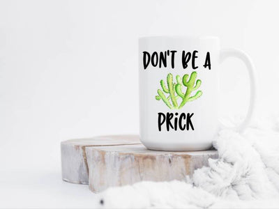 15 oz white ceramic coffee mug handle to the right Dont be a Prick in black with a green cactus in center mug is sitting on a white wash wood slab with a white blanket snuggled up to the wood and mug