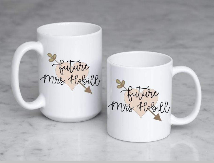Future Mrs. with last name in black text on top of peach heart with leafy arrow poking through in olive green displays both size mugs