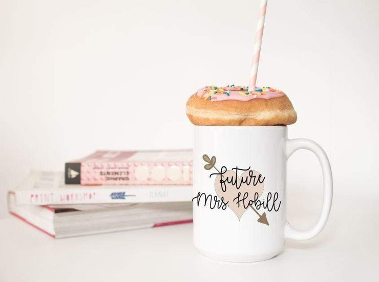 Future Mrs. with last name in black text on top of peach heart with leafy arrow poking through in olive green with a donut on top of mug and books behind it