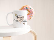 Future Mrs. with last name in black text on top of peach heart with leafy arrow poking through in olive green on edge of white table with donut hanging on side of mug with pink frosting and sprinkles